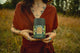 Person in a rust-red wrap dress holding The Steward herbal tea canister, labeled Deeply Nourishing, in a dry field
