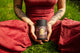 Person seated on grass in a red outfit holding The Lovers herbal tea canister labeled Rise To The Occasion